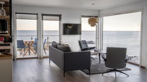 Ein modernes Wohnzimmer mit Sofa, Sessel und Fernseher, mit Blick auf eine malerische Küstenlandschaft.