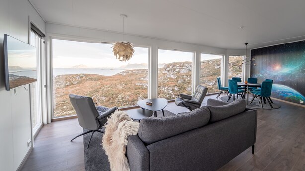 Ein großzügiger Wohnbereich mit einem großen Fenster, das einen schönen Ausblick auf Berge und ein Gewässer bietet. Es gibt ein graues Sofa, einen Esstisch mit Stühlen und eine hängende Deckenleuchte.