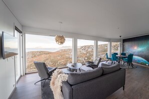 A spacious living area with a large window offering a scenic view of mountains and a body of water. There is a gray sofa, a dining table with chairs, and a hanging light fixture.