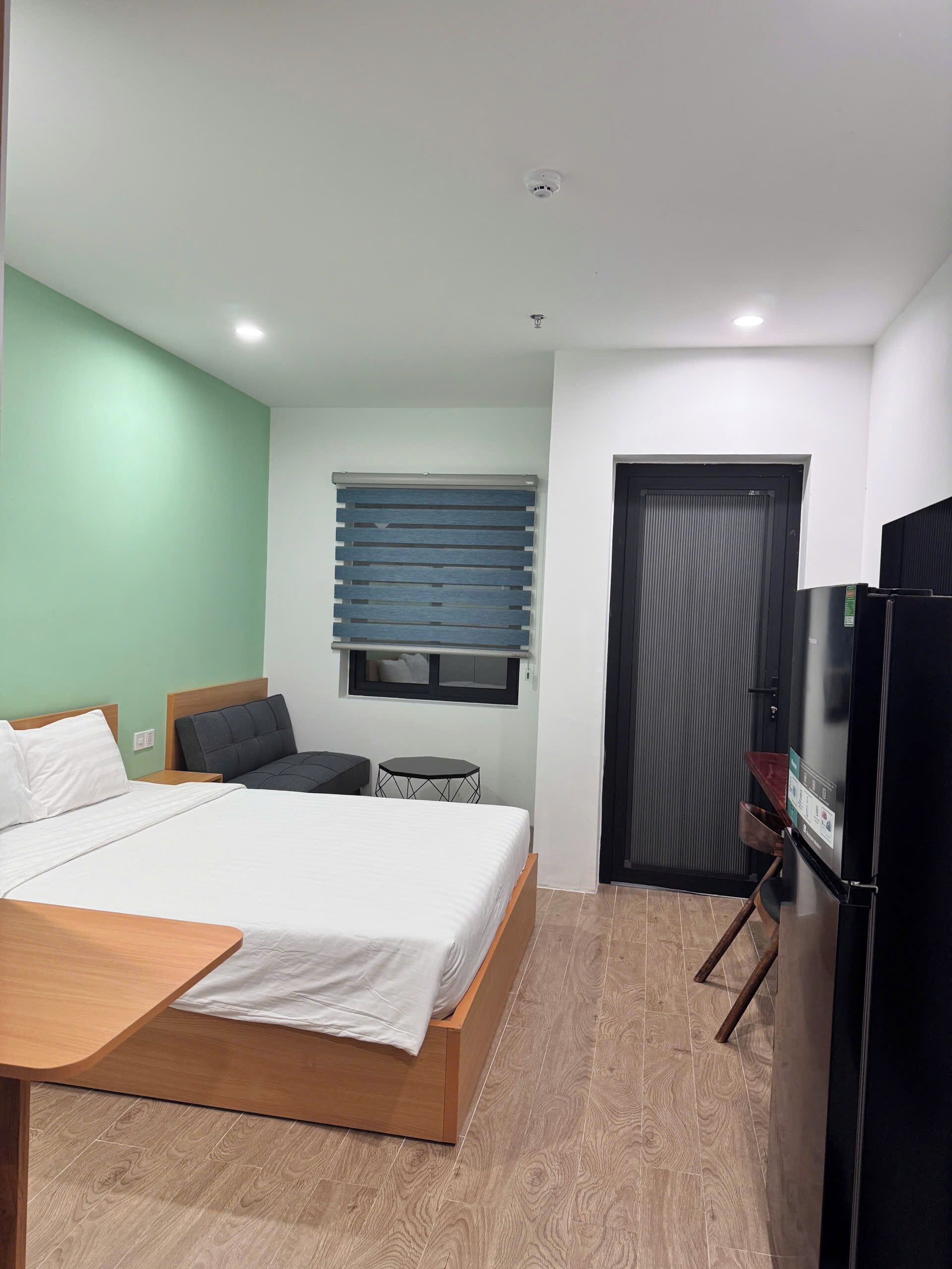 A bedroom with a bed, a small table, a chair, a window with blinds, a door, and a refrigerator.