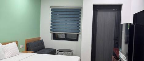A bedroom with a bed, a small table, a chair, a window with blinds, a door, and a refrigerator.