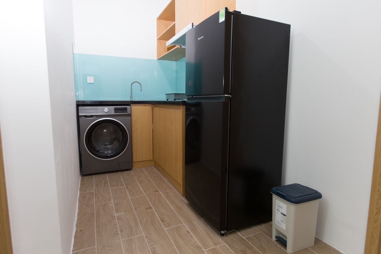 Eine kompakte Küche mit Waschmaschine, schwarzem Kühlschrank, Holzschränken und einem Mülleimer.