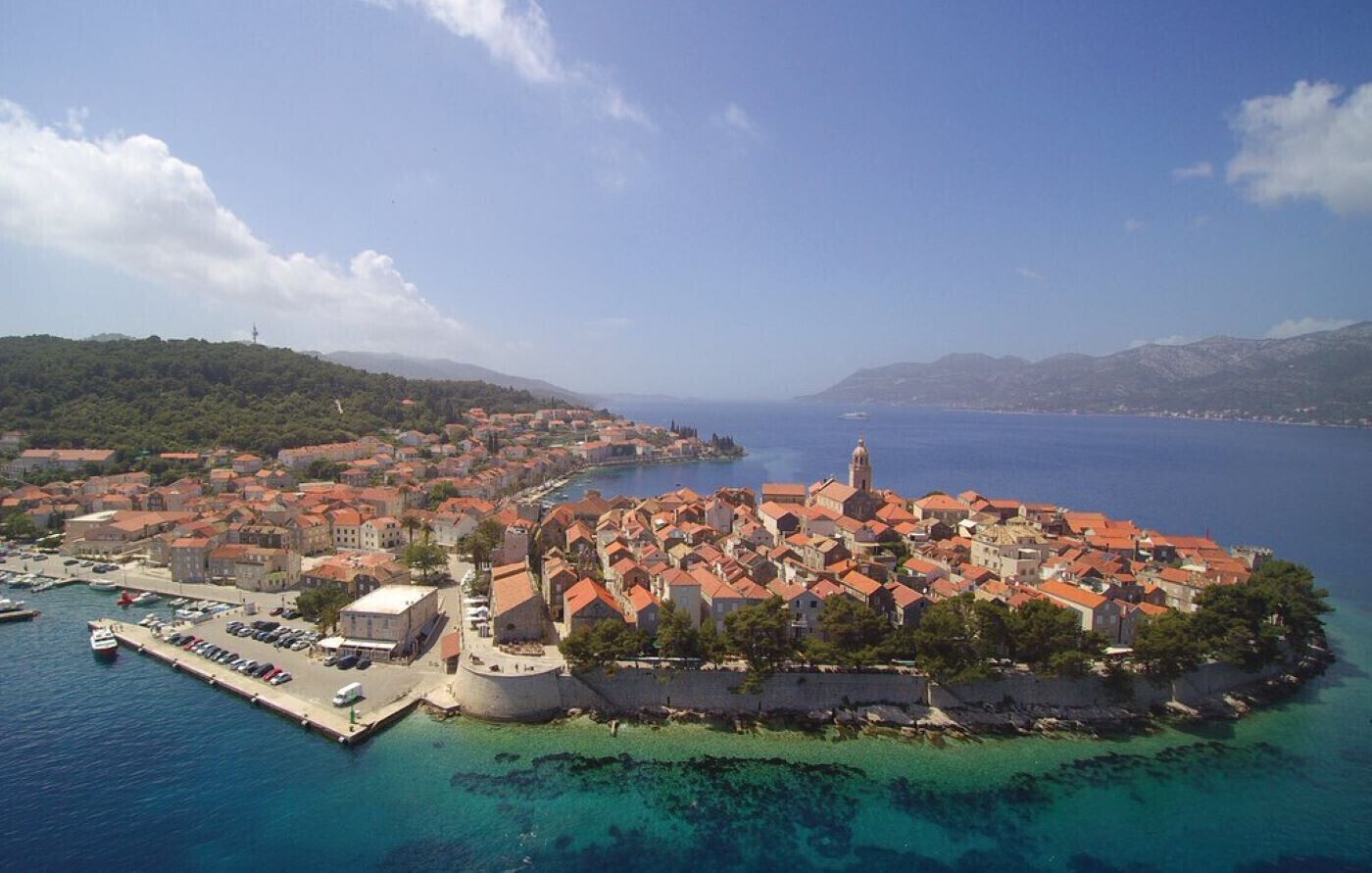 An aerial view of a coastal town with a historic fortification, clear blue waters, and a prominent church tower.