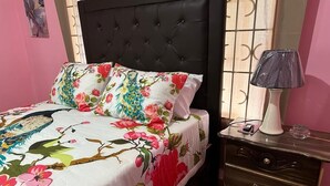A bed with a floral bedspread and a black headboard, a bedside table with a lamp, and a wall with a decorative panel. - Beautiful One Bedroom with WiFi in Brumalia Mandeville, Manchester (Mandeville)