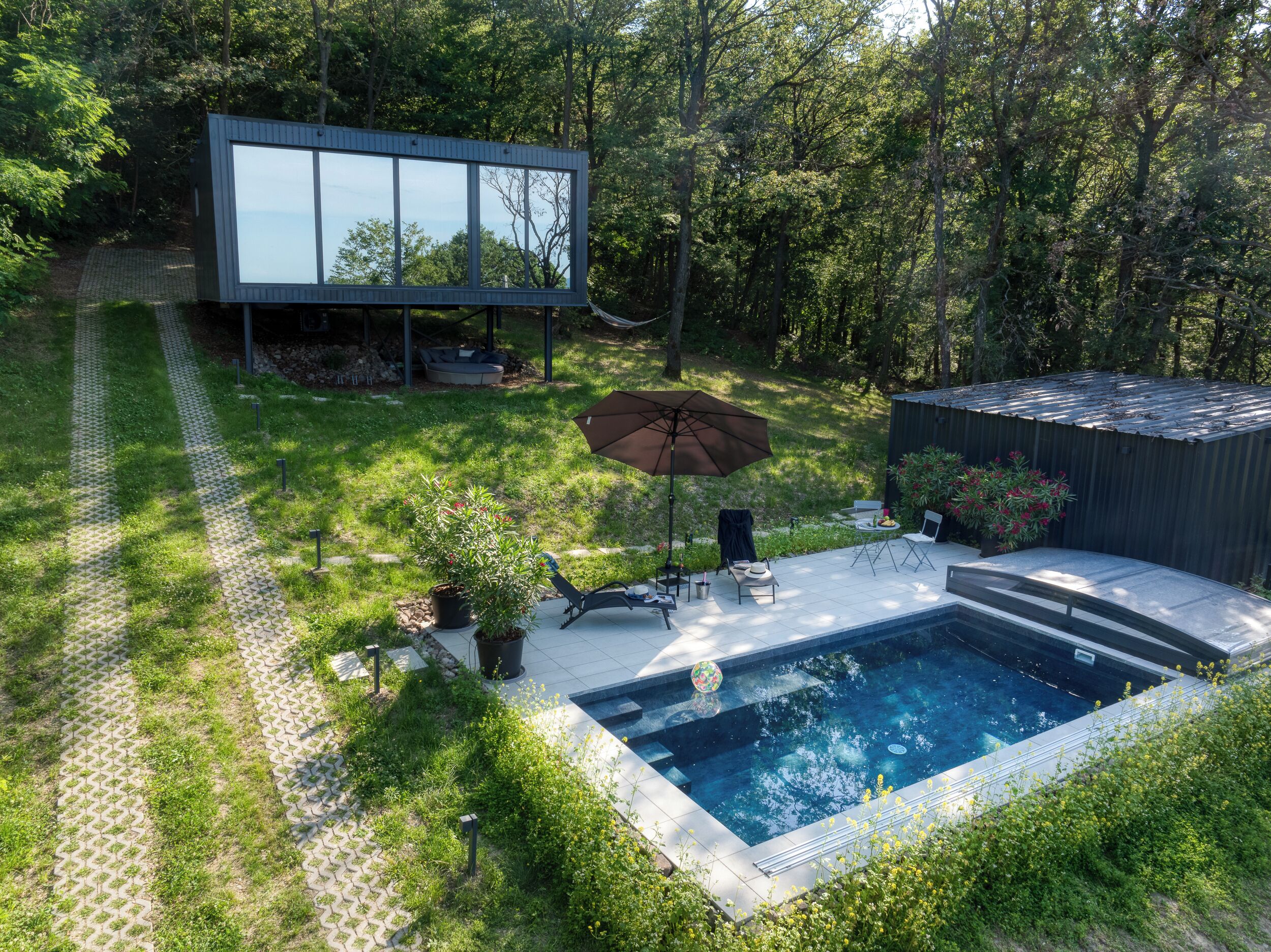 Zona de piscina exterior moderna amb una estructura de vidre, terrassa amb seients i un petit jardí.
