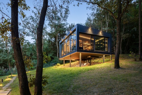 Une structure moderne en bois, surélevée, dotée de grandes fenêtres en verre et entourée d’arbres et d’une zone herbeuse.