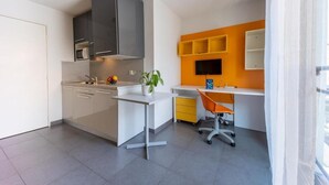 A compact kitchen with modern appliances, a dining area with a desk and chair, and a bright orange accent wall. - Le Campus Appartement (Nice)