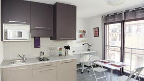 A modern kitchen with a microwave, sink, and cabinets. There is a white desk with chairs and a window with a view of buildings. - Le Campus Appartement (Nice)