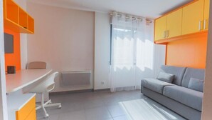 A compact living space with a kitchenette, a dining area, a sofa, and a window with sheer curtains. - Le Campus Appartement (Nice)