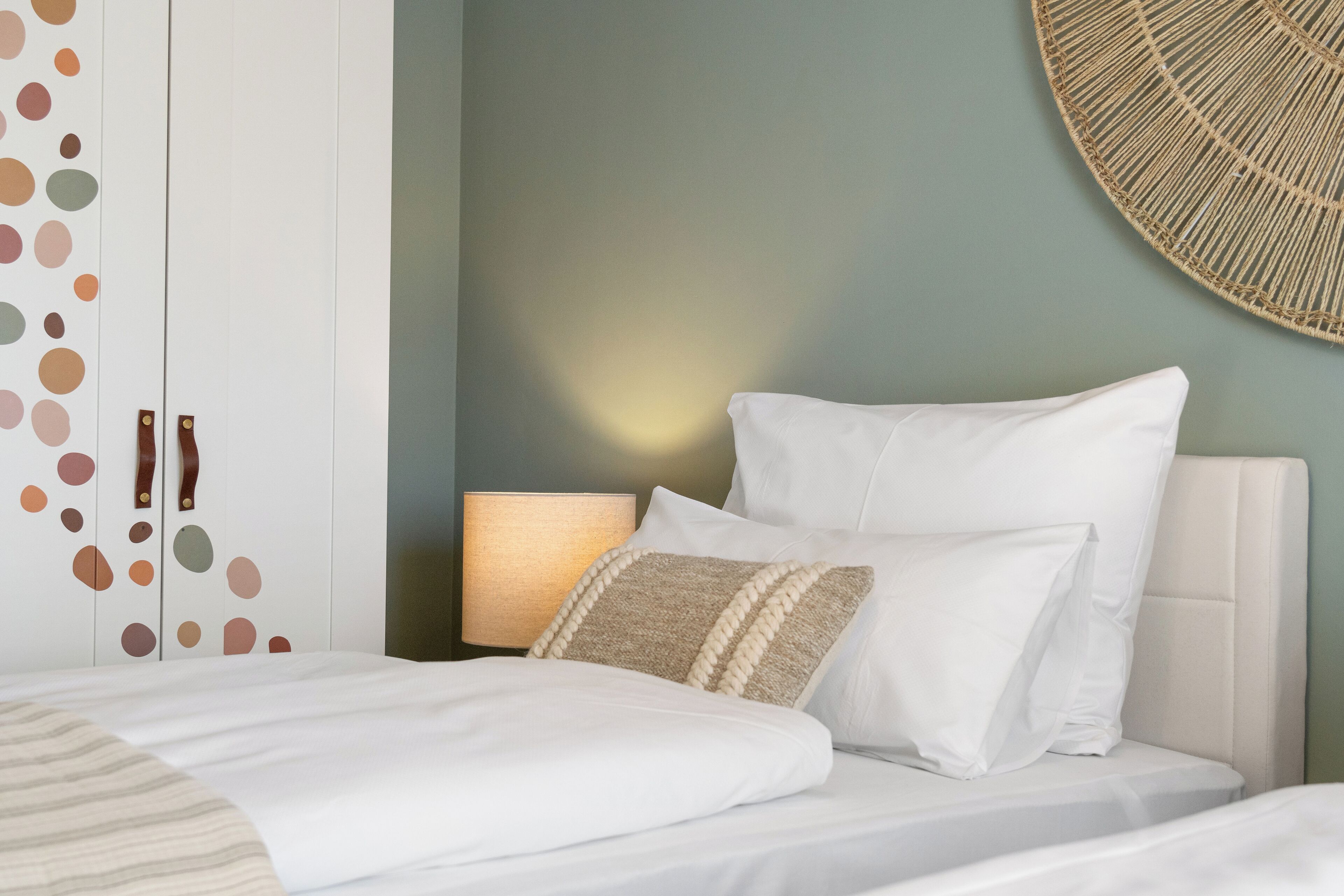 Een net opgemaakt bed met witte beddengoed en twee kussens, een bedlampje, en een wand met een rond geweven ontwerp.