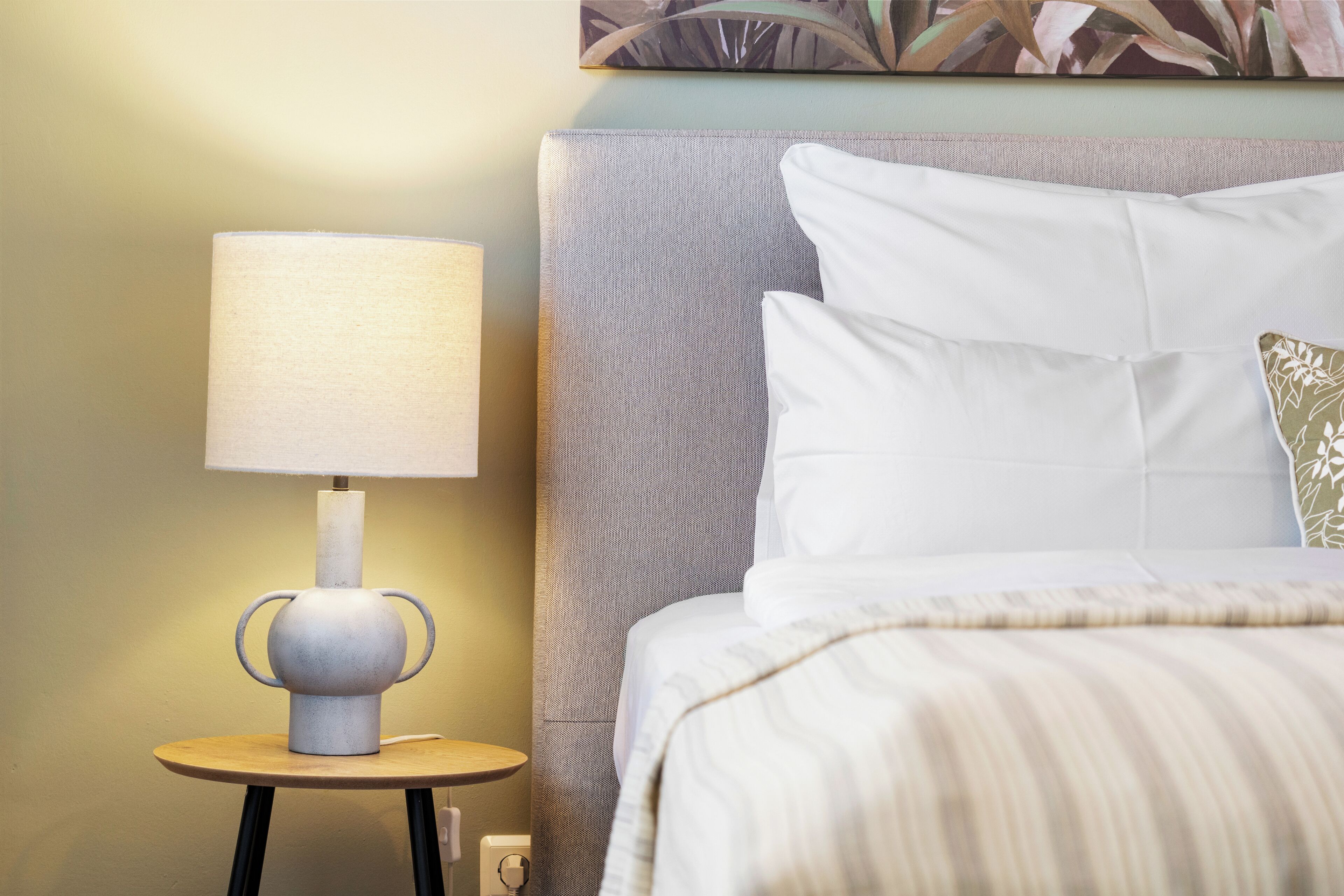 Un lit bien fait, avec du linge de lit blanc et une couverture à rayures, une lampe de chevet avec un abat-jour blanc, et une petite table ronde avec une lampe.