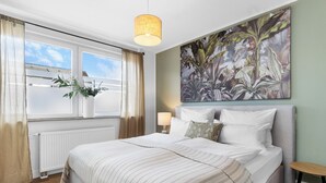 Un dormitorio con una cama grande, una ventana con cortinas, una planta colgante y un cuadro en la pared.