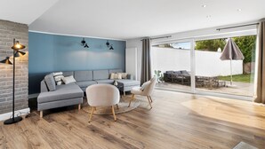 Una sala moderna con un sofá seccional gris, dos sillones, una mesa de centro y una lámpara de pie. Amplias ventanas ofrecen vistas al exterior.