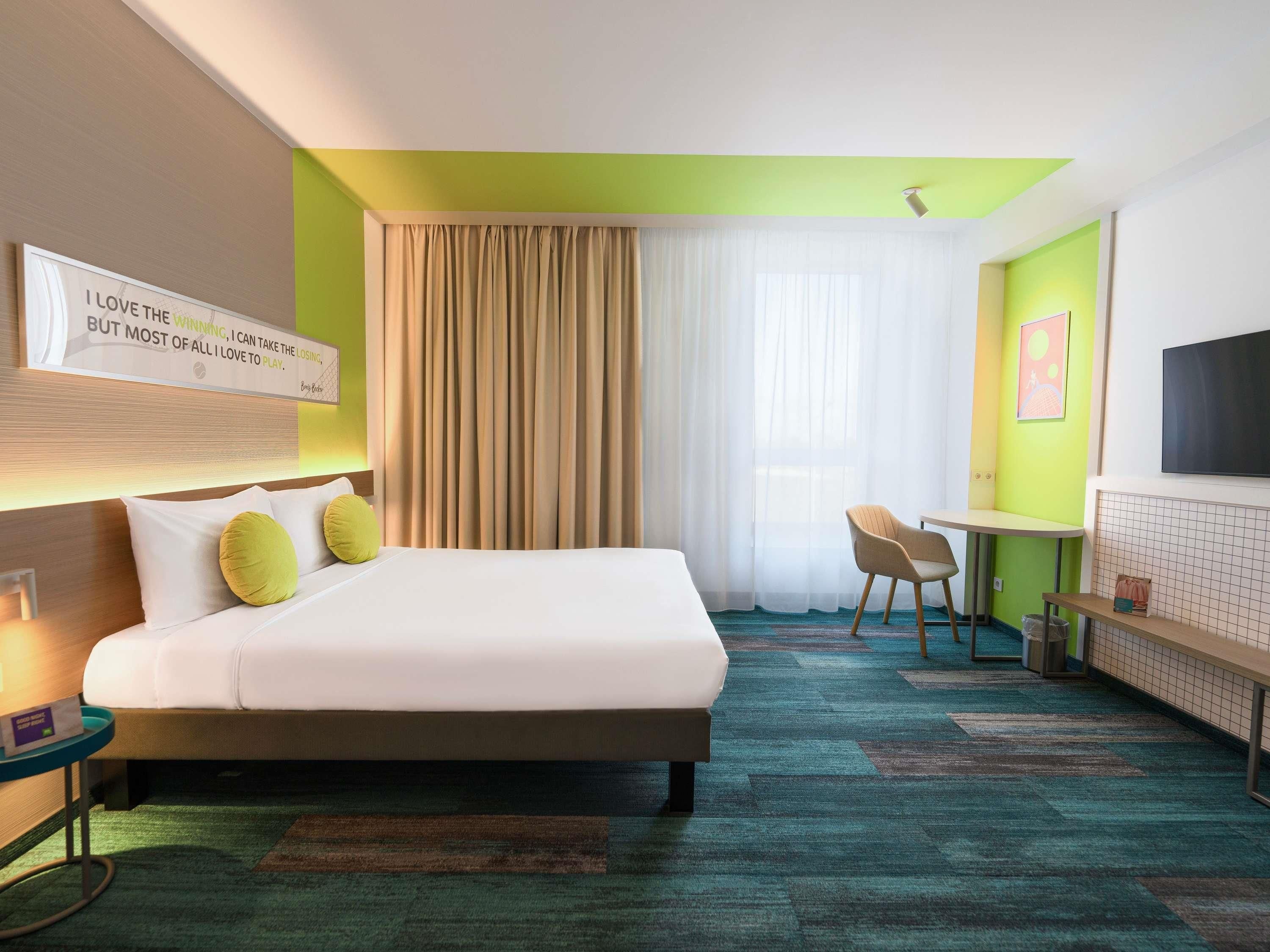 A hotel room with a large bed, a desk, a chair, a TV, and a window with curtains.