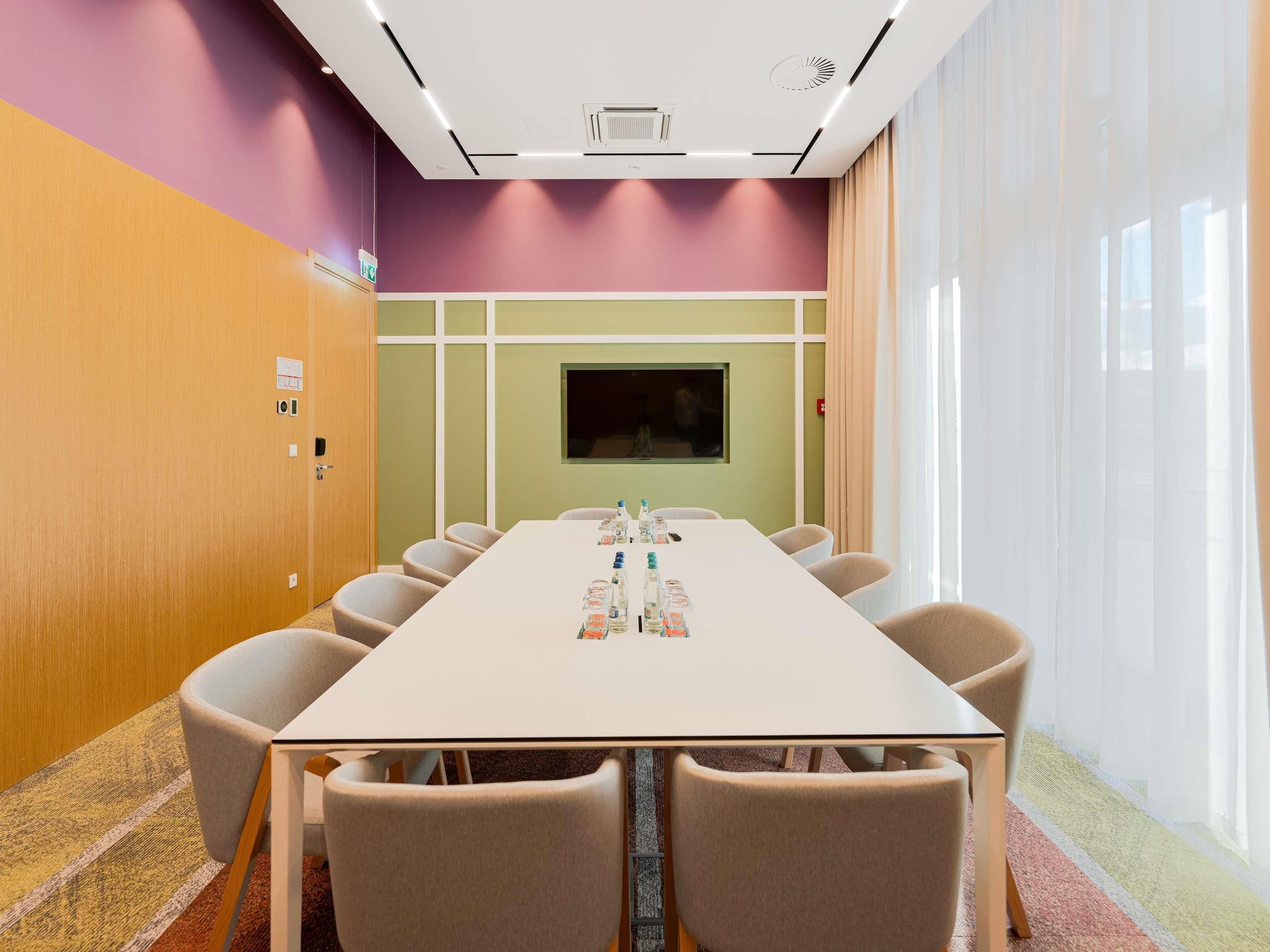 A conference room with a long table, chairs, a television screen, and a window with curtains.