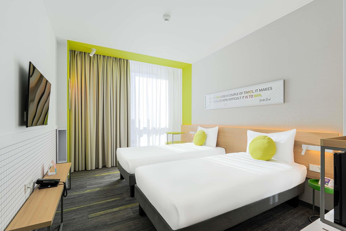 A hotel room with two beds, a desk, a chair, a TV, and a window with curtains.