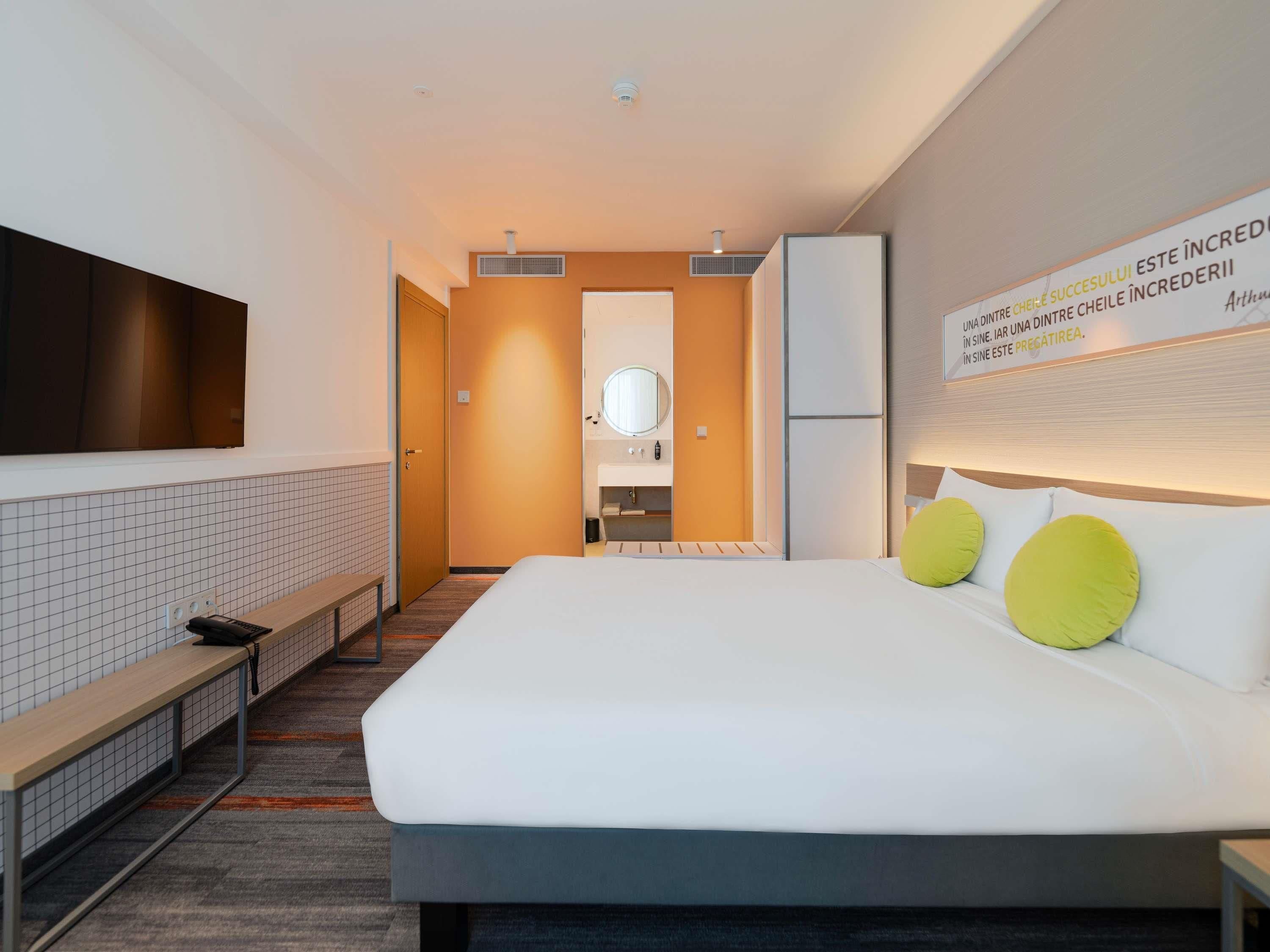 A modern hotel room with a bed, a bench, a mirror, a sink, and a television.