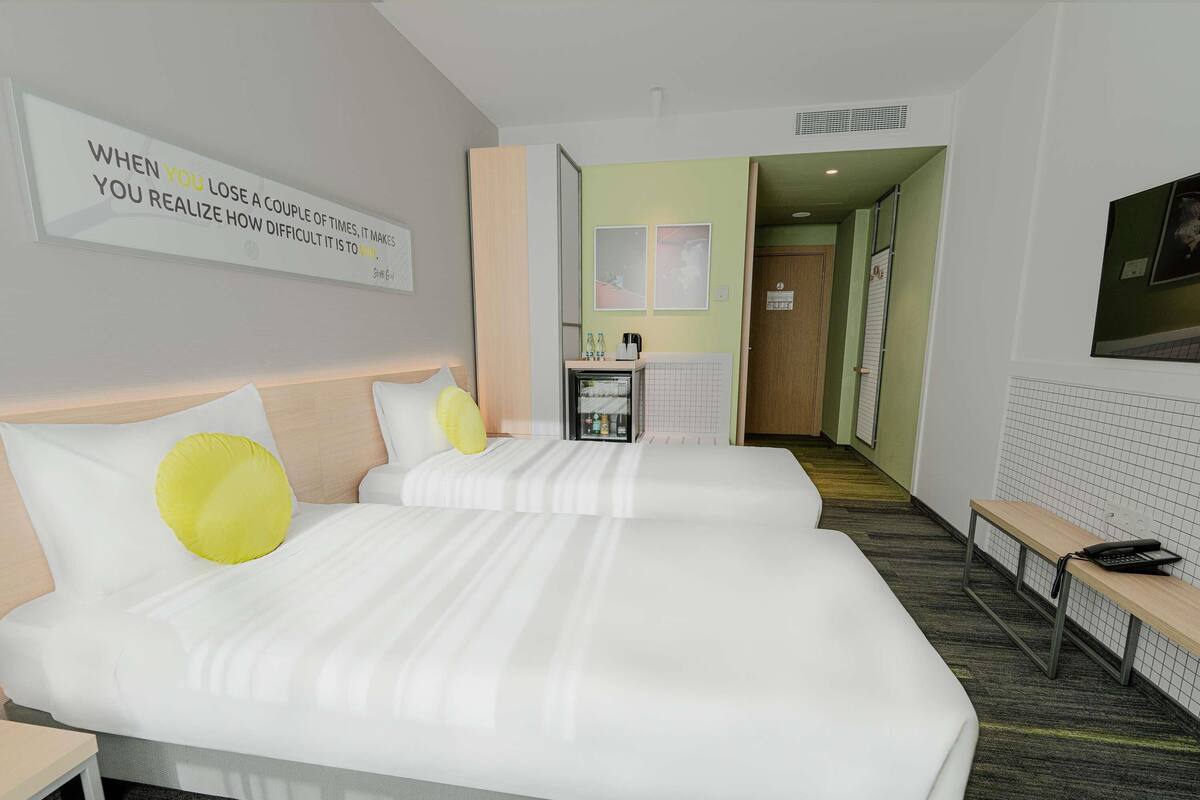 A hotel room with two beds, a bedside table, a desk, a chair, a small kitchenette, and a bathroom visible through an open door.