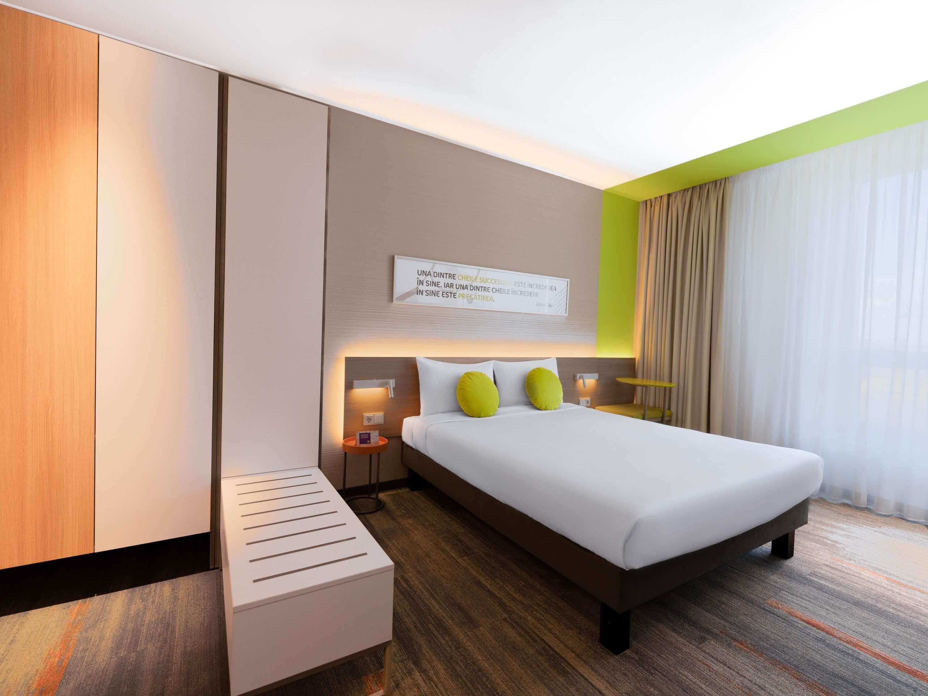 A modern hotel room with a bed, bedside tables, a nightstand, a wall-mounted light, and a window with curtains.