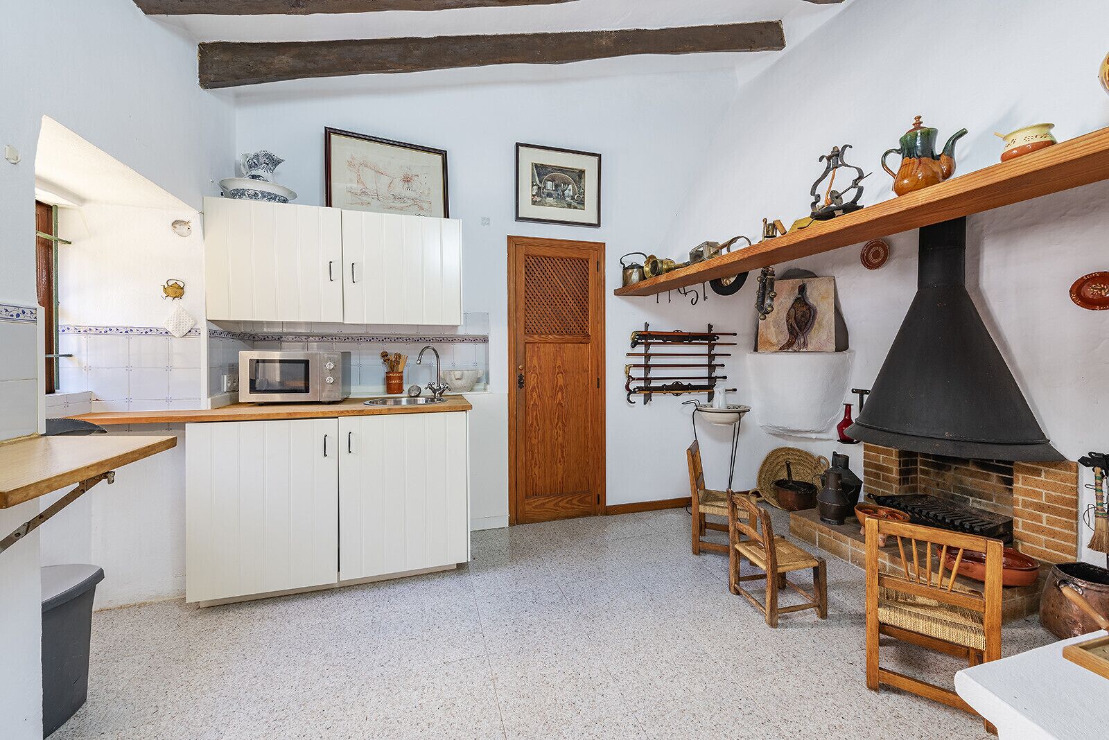 A kitchen with white cabinets, a microwave, a wooden shelf with kitchen utensils, a fireplace, and a door with a patterned design.
