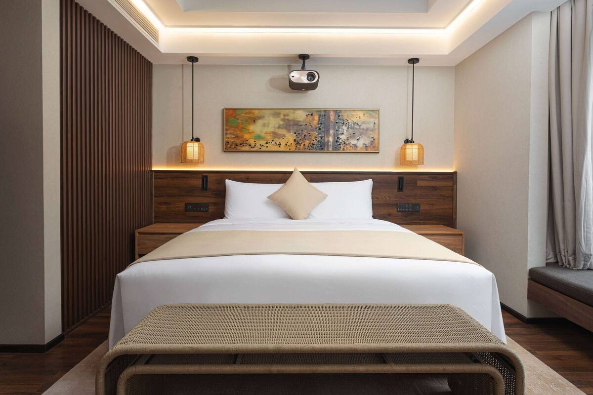 Una habitación de hotel moderna con una cama grande, cabecera de madera, dos mesitas de noche, un proyector montado en la pared y un cuadro abstracto de gran tamaño.