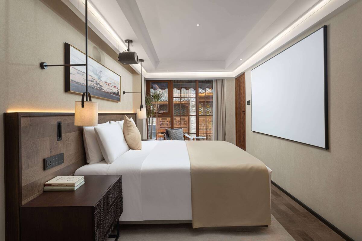 Una habitación de hotel moderna con una cama grande, mesita de noche y vistas de la ciudad a través de amplios ventanales.