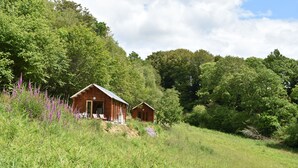 Eine rustikale Holzhütte, eingebettet in ein üppiges Grünfeld mit hohem Gras und violetten Blüten.