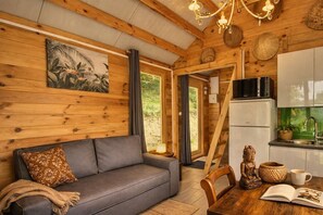 Interior acogedor de una cabaña con sofá, mesa de comedor, zona de cocina y un amplio ventanal.