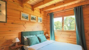 Ein Zimmer in einer Holzhütte mit einem Bett, einem Nachttisch, einer Lampe und einem Fenster mit Vorhängen.