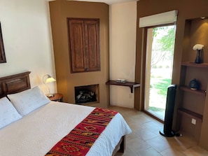 A bedroom with a bed, a nightstand, a lamp, a fireplace, a window, and a shelf with decorative items. - Serene Oasis in San Miguel de Allende 6-bedroom Villa With Hot Tub (San Miguel de Allende)