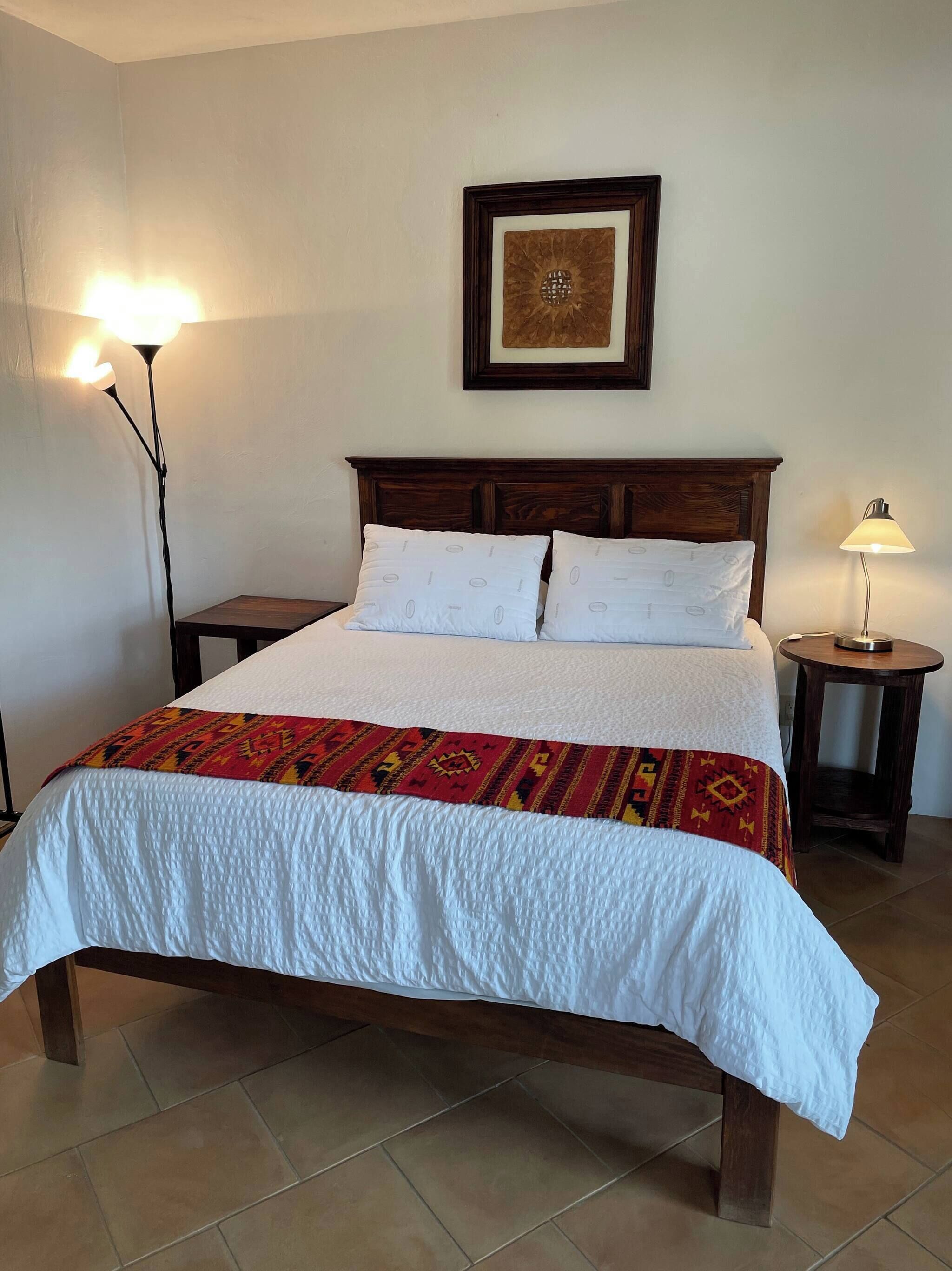 Un lit bien fait, recouvert d’une couvre-lit à motifs, deux lampes de chevet, un tableau encadré accroché au mur et une petite table de chevet en bois.