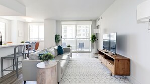 Ein modernes Wohnzimmer mit Sofa, Couchtisch und TV-Schrank. Es gibt einen Essbereich mit Stühlen und Tisch sowie einen Balkon mit Blick auf Gebäude.