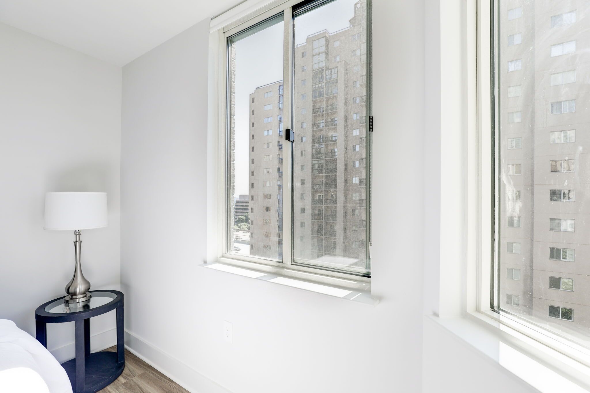 Una camera d'albergo con una grande finestra, un comodino con una lampada e vista su edifici alti.