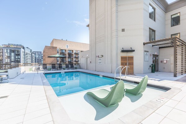 A rooftop pool with a clear blue pool, green lounge chairs, and a modern building with a brick facade.
