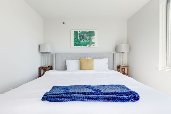A neatly made bed with a blue blanket, white bedding, and a green abstract painting above it.