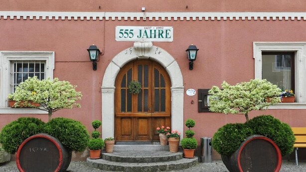 A building with a sign reading "555 JAHRE" and a wooden door with a wreath. There are potted plants and barrels on the steps. - Hotel Grüner Baum (Pommersfelden)
