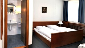 A hotel room with a wooden bed, a bedside table with a lamp, a window with curtains, and a bathroom with a sink and mirror. - Hotel Grüner Baum (Pommersfelden)