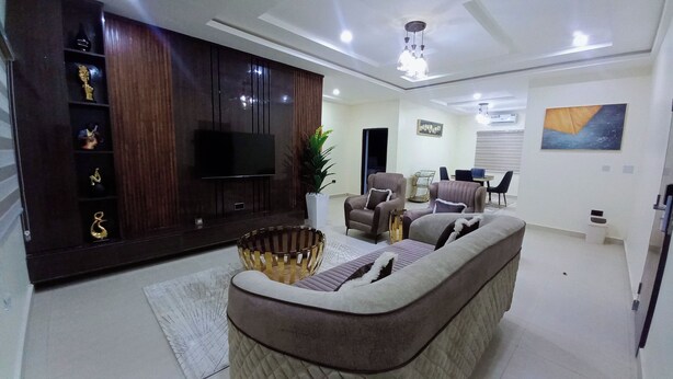 A modern living room with a flat-screen TV, a sofa, a coffee table, and a dining area with chairs and a table. - Kelvic Systems Apartments (Abuja)