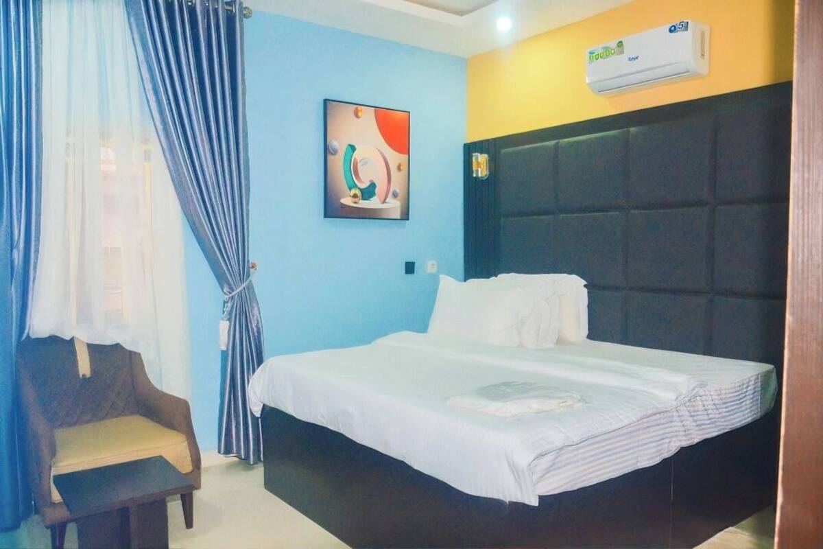 A hotel room with a bed, a chair, a wall-mounted air conditioner, and a painting.