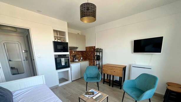 A compact living space with a bed, a small kitchenette, a dining area, and two teal chairs. - L'Escale - Studio with loggia in the city center (Courseulles-sur-Mer)