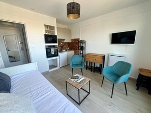 A compact living space with a bed, a small kitchenette, a dining area, and two teal chairs. - L'Escale - Studio with a Loggia in the City Center (Courseulles-sur-Mer)