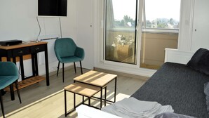 A modern, well-lit room with a bed, a desk with a computer, a teal chair, and a view of the city through a large window. - L'Escale - Studio with loggia in the city center (Courseulles-sur-Mer)