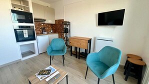 A compact living space with a kitchenette, two teal chairs, a coffee table, a TV, and a small stool. - L'Escale - Studio with loggia in the city center (Courseulles-sur-Mer)