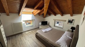 Une chambre avec un plafond en bois, un lit recouvert de draps blancs, un téléviseur posé sur une table de chevet et une fenêtre donnant sur des immeubles.