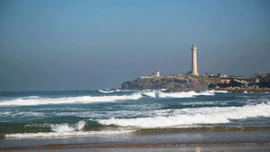 A coastal scene with a lighthouse on a rocky outcrop, waves crashing onto the shore, and a clear sky. - Stay'Inn - Rafina Sunset (Casablanca)