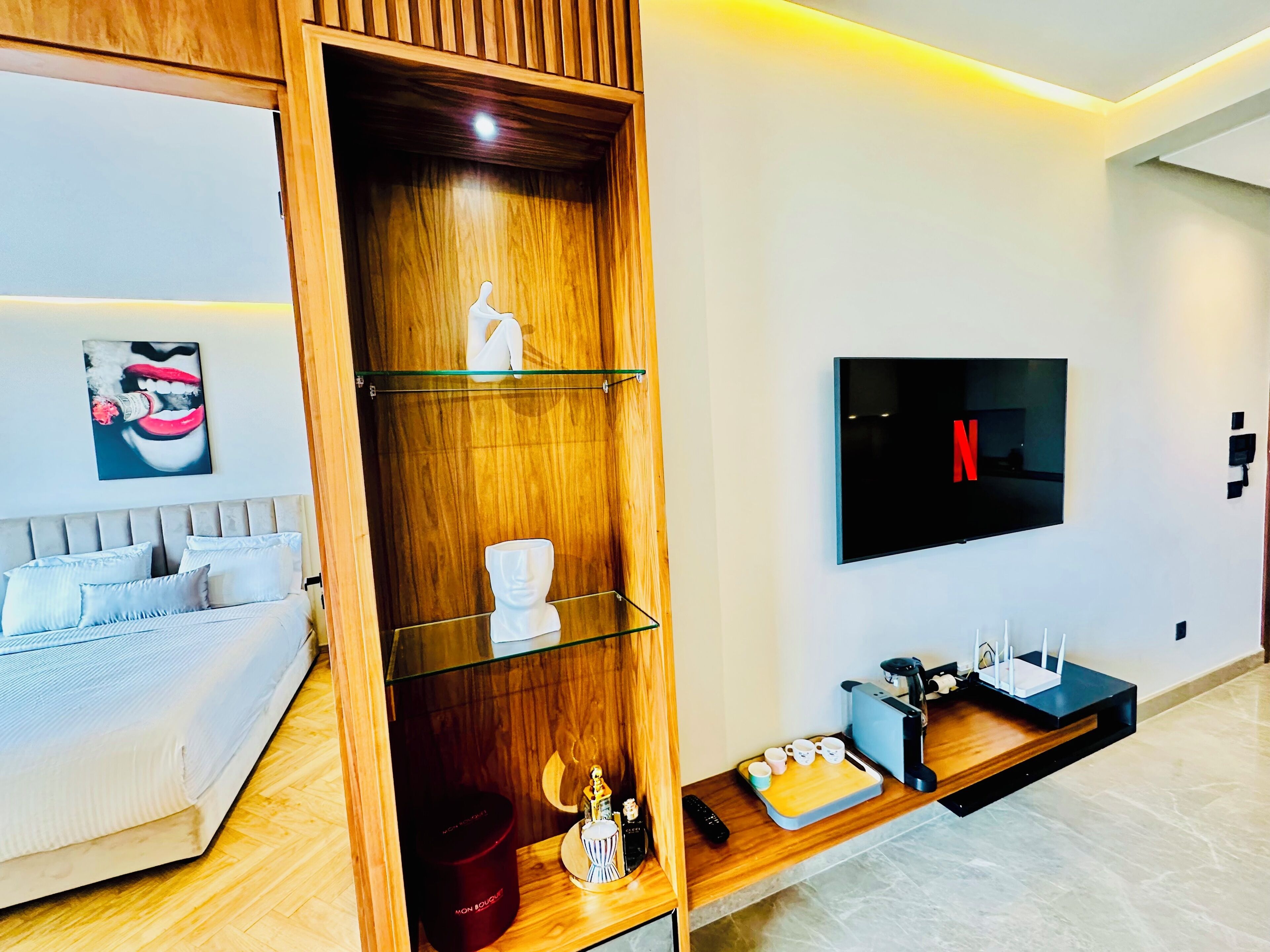 A modern living room with a grey sofa, a wooden cabinet with glass shelves, a mounted TV, and a coffee station.