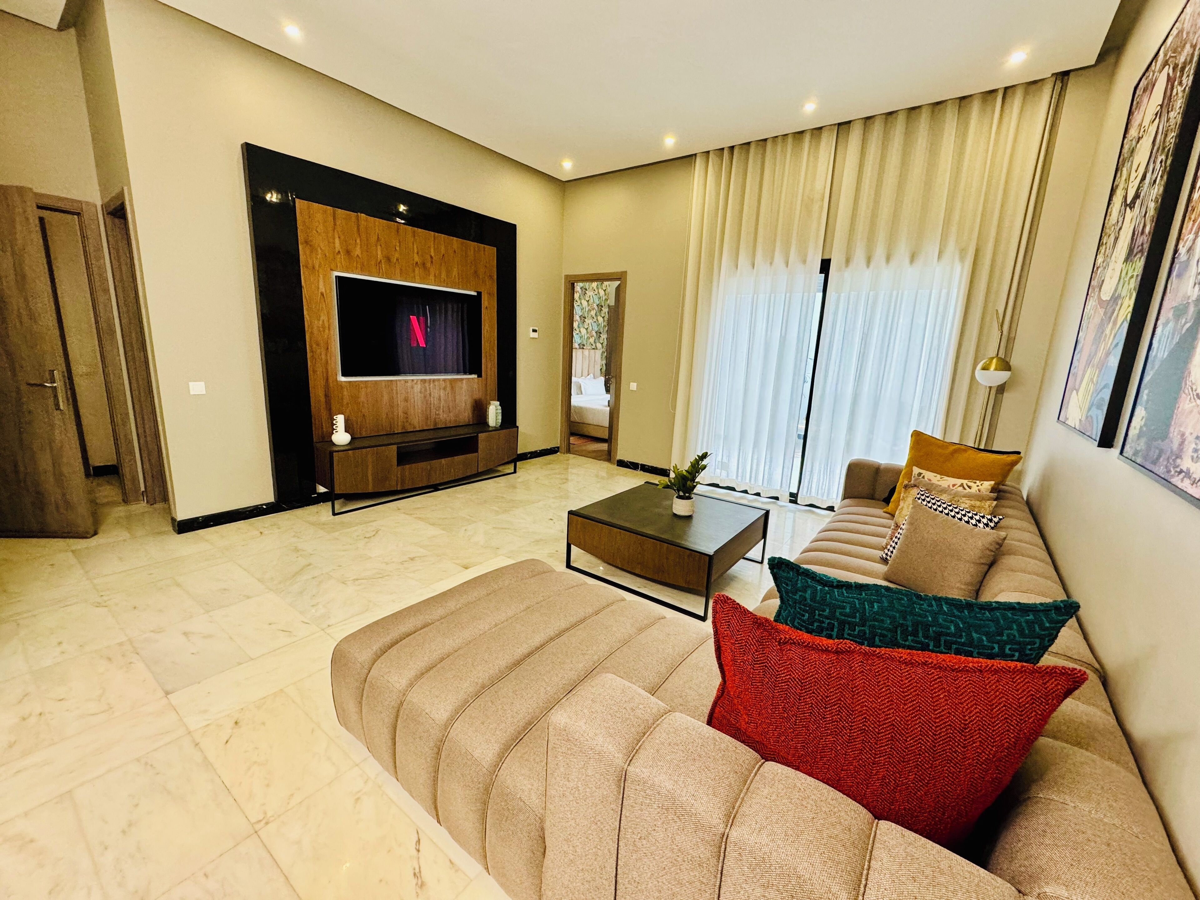 A modern living room with a flat-screen TV, a sofa with patterned cushions, a coffee table, and a large window with curtains.