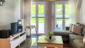 A modern living room with a flat-screen TV, a sofa, a coffee table with a plant, and large windows with a view of greenery outside.