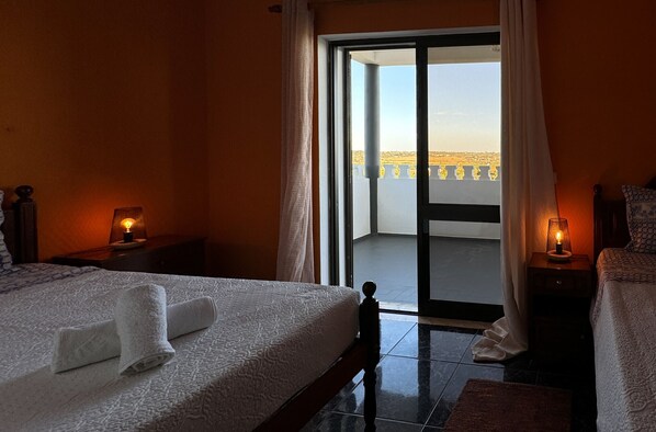 A hotel room with a bed, two bedside tables with lamps, a balcony with a view, and a sliding glass door.
