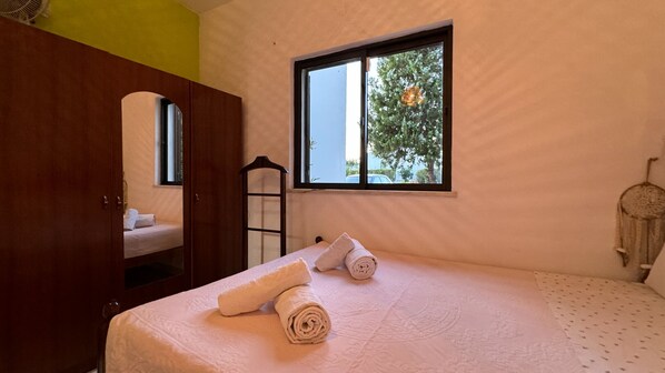A bedroom with a bed, a window with a view of trees, a wardrobe, and a bedside table.